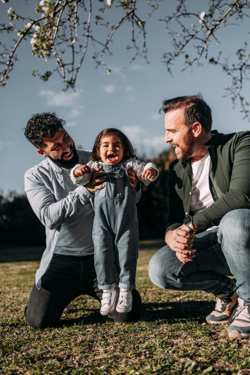 Male gay couple holding their little daughter in the air who laughs happily. 